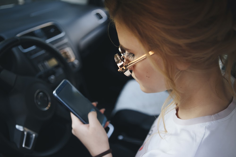 Photograph of a teen sitting at the wheel of a car, looking at a cellphone. 